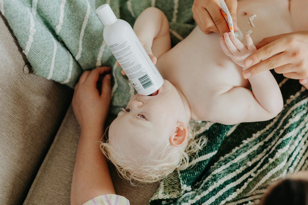 A cute baby lies comfortably while holding a lotion bottle during a skincare routine.