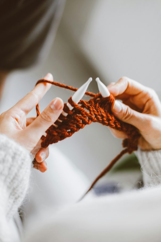 Detailed close-up of hands knitting a red yarn indoors, showcasing a cozy hobby.