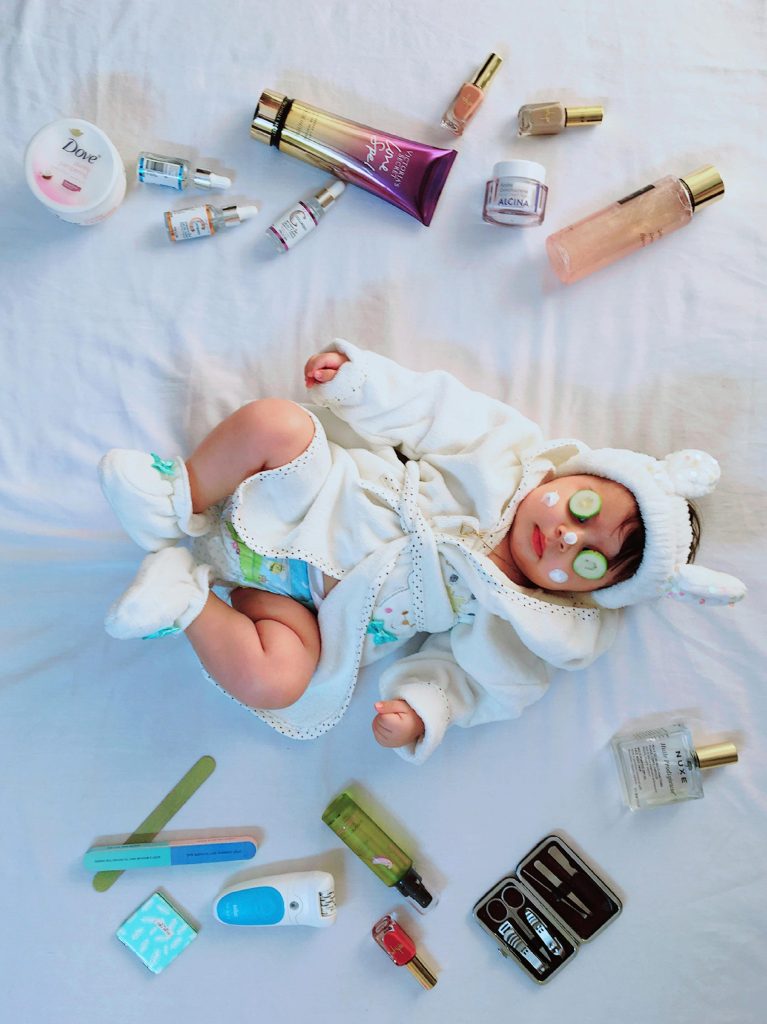 Cute baby with cucumber eye mask surrounded by skincare items on a bed.